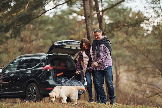 Un couple d'âge mûr heureux se promène avec son chien dans la forêt d'automne ou de printemps près d'une voiture moderne