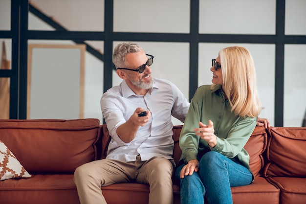 Couple d'âge mûr à lunettes assis sur le canapé et allumer la télévision