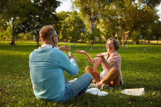 Un couple d'aînés actifs et ludiques soufflant des bulles de savon pendant qu'ils se reposent dans le parc.