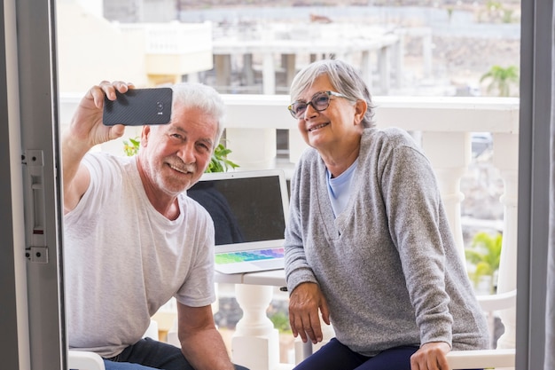 Couple d'aînés sur la terrasse de leur maison avec ordinateur portable et assis en train de prendre un selfie ensemble - femme et homme retraités mûrs et détendues prenant une photo