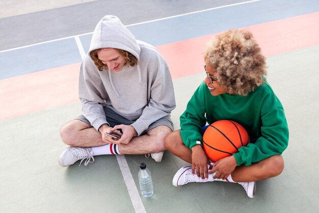 Couple d'amis discutant et partageant un bon moment assis sur le terrain après un entraînement de basket-ball dans un parc de la ville concept d'amitié et de sport urbain dans la rue