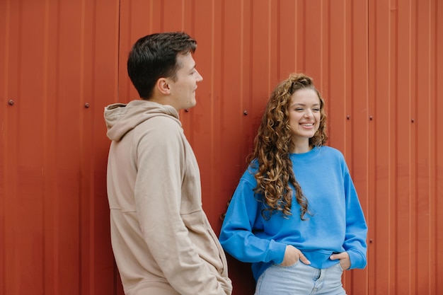 Un couple d'amis, un garçon et une fille, parlent sur le fond d'un mur rouge à l'extérieur.