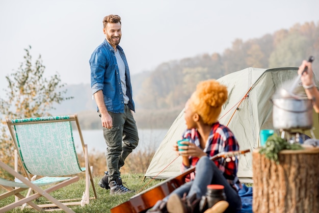 Couple d'amis multiethniques habillés avec désinvolture parlant pendant les loisirs de plein air avec tente près du lac