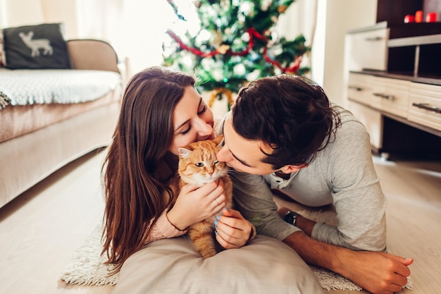 Couple amoureux allongé près de l'arbre de Noël et jouant avec un chat à la maison. Heureux homme et femme relaxant