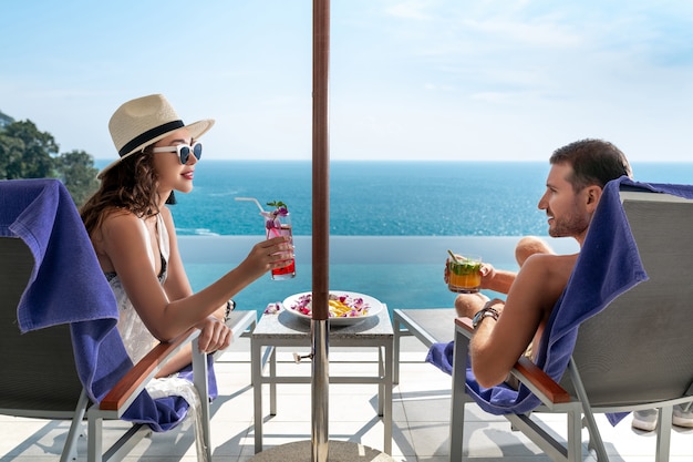 Couple d'amoureux dans un complexe tropical. Homme et femme se regardent assis sur des transats au bord de la piscine et dégustent des cocktails.