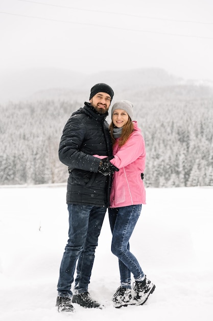 Couple d'amoureux en doudoune lors d'une promenade hivernale. Homme et femme posant dans la forêt givrée. Rendez-vous romantique en hiver. Ambiance de Noël d'une jeune famille. Histoire d'amour d'hiver