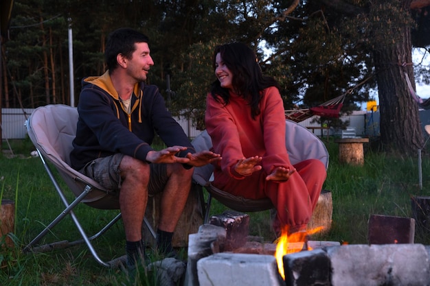 Un couple amoureux est assis près du feu de camp en plein air dans la cour de la maison sur des chaises de camping une soirée romantique un homme et une femme réchauffent leurs mains près du feu boivent du thé