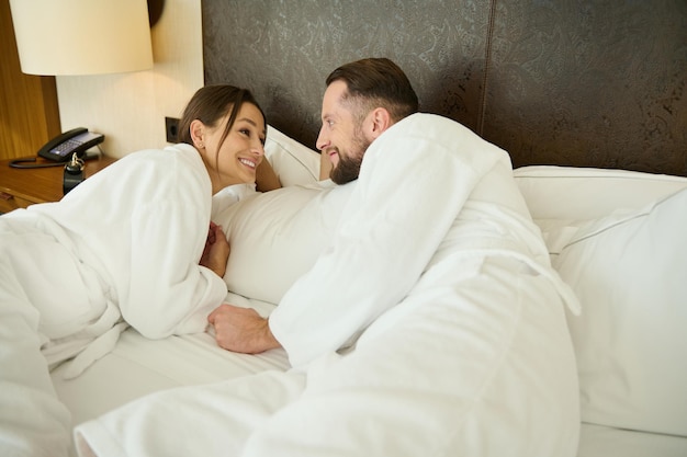 Couple amoureux, jeunes mariés en lune de miel. Belle femme portant un peignoir en éponge blanche allongée dans son lit avec son mari et profitant de l'intimité du week-end ensemble à la maison. Relation familiale