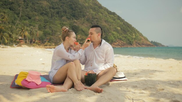 Le couple d'amoureux joyeux tenant et mangeant des tranches de pastèque sur la mer de plage de sable tropical