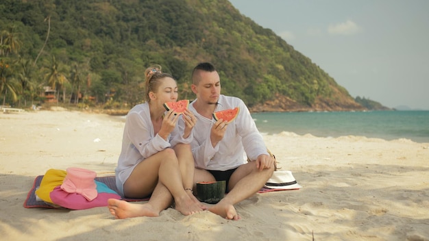 Le couple d'amoureux joyeux tenant et mangeant des tranches de pastèque sur la mer de plage de sable tropical