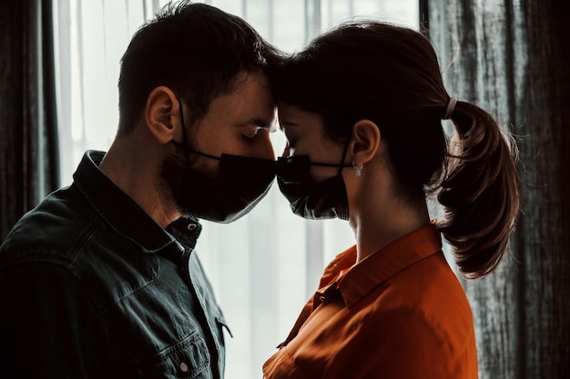 Couple amoureux de masques faciaux debout à côté de la fenêtre et se regardant