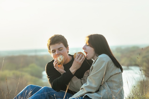 Un couple amoureux, un mec et une fille dans la nature aux rayons du soleil couchant se nourrissent de hamburgers