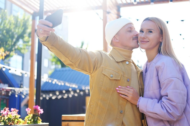 Un couple amoureux un mec et une fille prennent un selfie ensemble par temps ensoleillé Jeune homme embrassant une fille sur la joue