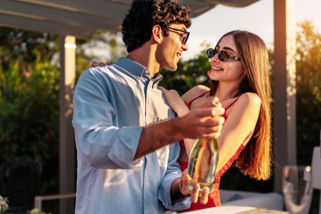 Un couple d'amoureux ouvre une bouteille de champagne à côté d'amis assis à table pour célébrer la fête de fiançailles