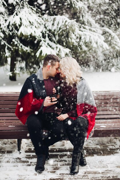 Couple amoureux petit ami et petite amie posant et assis sur un banc dans la forêt d'hiver. Homme assez blond et souriant regardant la caméra et tenant des tasses avec du café dans les mains. Notion d'amour.