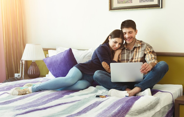 Couple d'amoureux planifiant un voyage de noces sur un ordinateur portable assis sur le lit dans une chambre d'hôtel