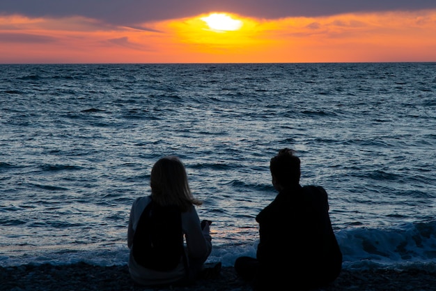 Couple amoureux regardant le coucher du soleil ensemble sur les vacances d'été de voyage à la plage. Silhouette de personnes par derrière assis en profitant de la vue sur la mer au coucher du soleil lors de vacances à destination tropicale. Couple romantique sur la plage