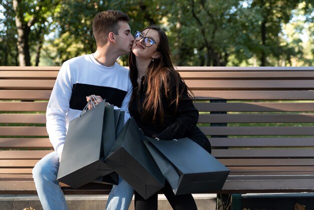 Couple d'amoureux reposant sur un banc dans le parc avec des sacs en papier après un shopping réussi Belle date