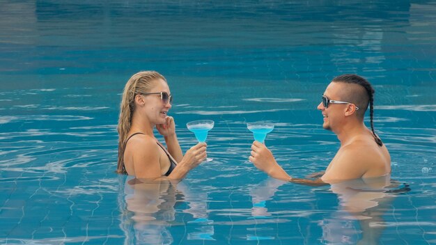 Le couple d'amoureux s'embrasse et s'embrasse, boit de l'alcool à cocktail bleu dans la piscine de l'hôtel en plein air. Portrait d'un homme et d'une femme caucasiens.