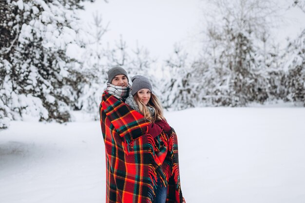 Couple amoureux se cachant avec une couverture dans une belle forêt d'hiver enneigée