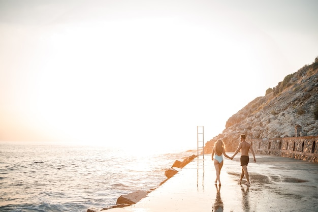 Un couple d'amoureux se promène le long de la plage au bord de la mer. Jeune famille au coucher du soleil au bord de la mer Méditerranée. Notion de vacances. Une femme en maillot de bain et un homme en short au coucher du soleil au bord de la mer. Mise au point sélective.