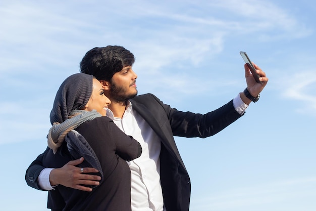 Couple arabe amoureux prenant selfie. Femme avec tête couverte et maquillage lumineux et homme en costume s'imaginant avec un téléphone portable. Amour, concept d'affection