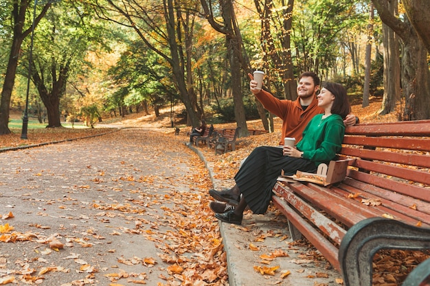 Photo couple assis au banc de parc public à la journée ensoleillée d'automne pointant vers l'espace de copie. boire pour aller