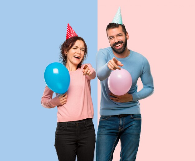 Couple avec des ballons et des chapeaux d&#39;anniversaire pointant du doigt quelqu&#39;un et riant beaucoup