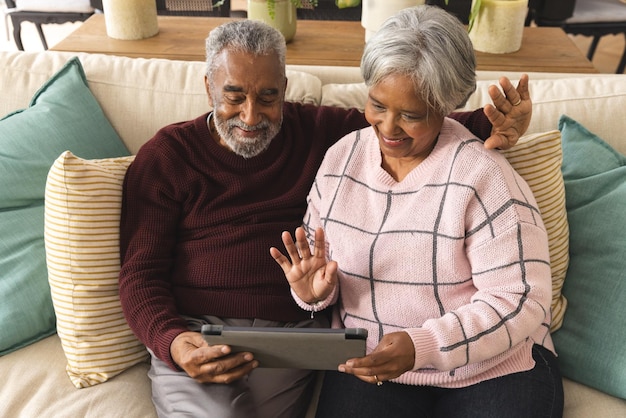 Un couple biracial âgé heureux assis sur le canapé et ayant un appel vidéo sur tablette à la maison