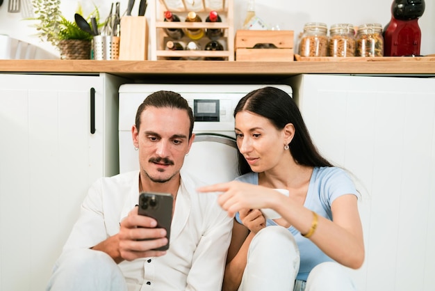 Un couple blanc heureux assis ensemble sur le sol de la cuisine pendant qu'un homme utilise un smartphone pour les médias sociaux ou le commerce électronique avec discussion Un jeune homme et une jeune femme discutent à la maison