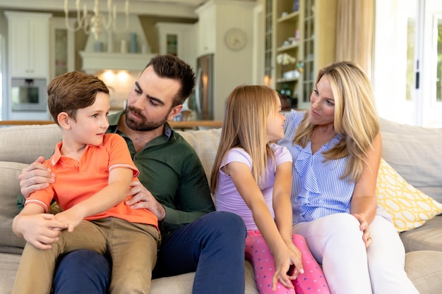 Un couple caucasien à la maison dans le salon assis sur le canapé, le père parlant à leur fils, assis sur ses genoux, et la mère parlant à leur fille, assise entre ses parents