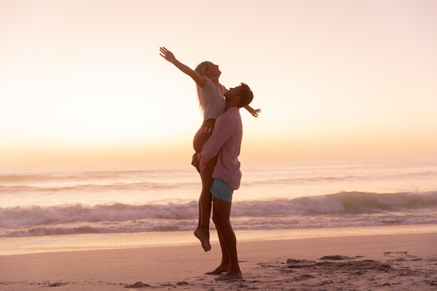 Couple caucasien profitant du temps à la plage, un homme soulève une femme, une femme élargit ses bras