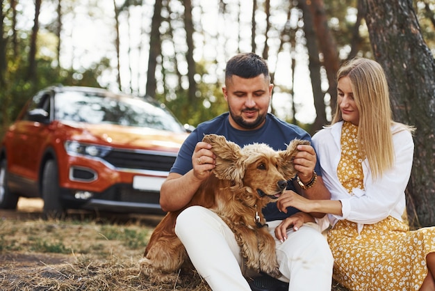 Un couple avec un chien passe un week-end à l'extérieur dans la forêt avec une voiture derrière eux.