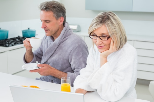 Couple dans des peignoirs avec du café et du jus en utilisant un ordinateur portable