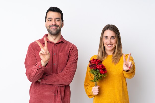 Couple dans la Saint-Valentin tenant des fleurs sur un mur isolé, souriant et montrant le signe de la victoire