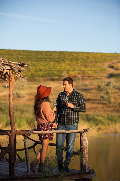 Couple dégustant un verre de vin sur un ponton en bois avec lac derrière lui au milieu d'un grand vignoble.