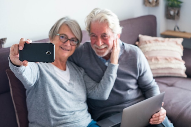 Couple de deux personnes âgées ou matures sur le canapé avec leur ordinateur portable et prenant un selfie avec son téléphone - personnes âgées de race blanche