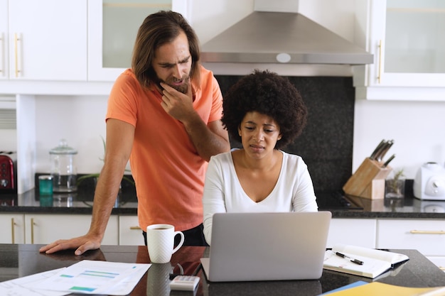 Couple diversifié assis dans la cuisine utilisant un ordinateur portable et payant des factures