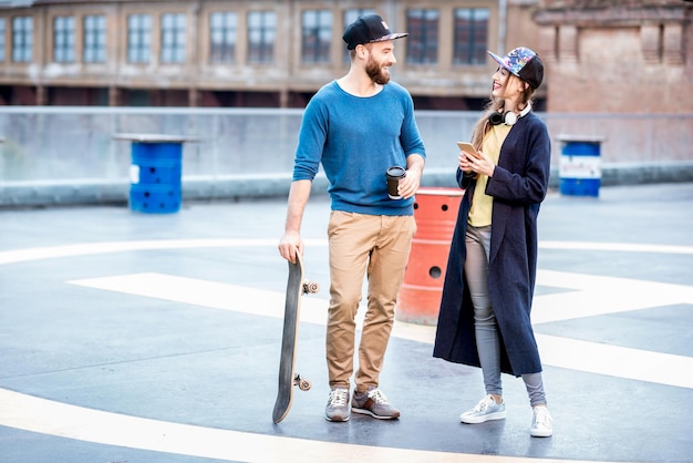 Couple élégant en chandails bleus et casquettes parlant ensemble debout avec une planche à roulettes à l'extérieur sur l'aire de jeux sur le toit