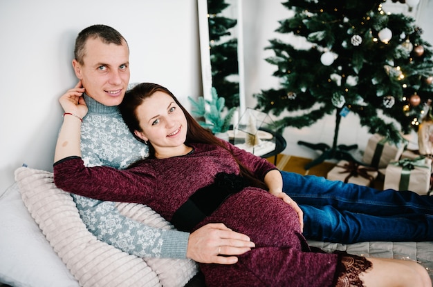 Couple enceinte allongé sur le lit près de l'arbre de Noël. Bonne année et joyeux Noël. Intérieur décoré de Noël. Concept de grossesse, vacances en famille, personnes et attente.