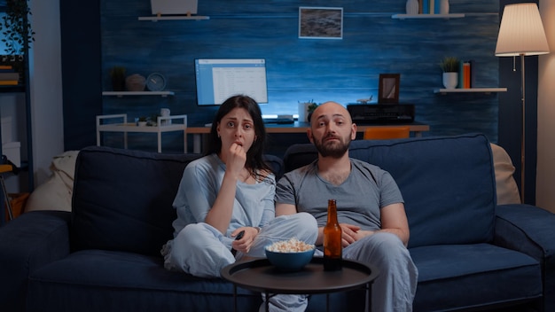 Couple étonné regardant un film à la télévision la nuit et mangeant du pop-corn, buvant de la bière en discutant sur des séquences de films. Femme étonnée concentrée et concentrée assise à la maison sur un canapé confortable, expression faciale