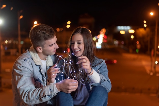 Couple d'étudiants amoureux sont assis sur fond de ville du soir avec une guirlande dans leurs mains