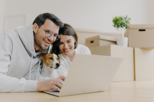 Un couple familial ravi rit en regardant des magasins en ligne sur un ordinateur portable moderne avec leur chien faire une pause après avoir déballé des boîtes en carton rénovation domiciliaire étant de bonne humeur Maison nouvellement louée