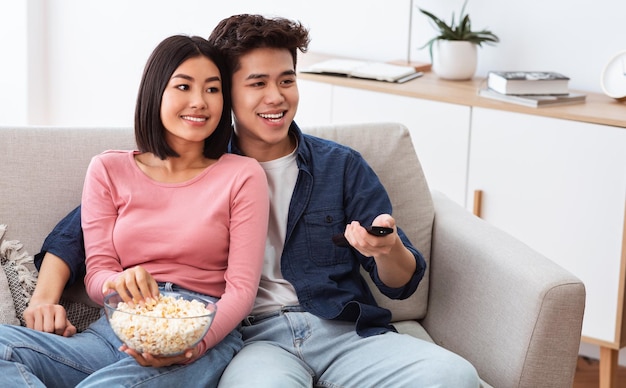 Couple de famille chinoise regardant la télévision ensemble et mangeant du pop-corn le week-end, changeant de chaîne de télévision avec télécommande assis sur un canapé dans le salon à la maison.