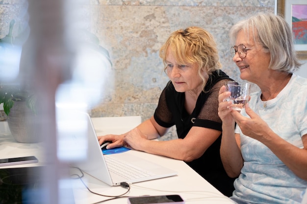 Photo couple de femmes mûres assises ensemble à la maison, naviguant sur un ordinateur portable, passant du temps à regarder les médias sociaux