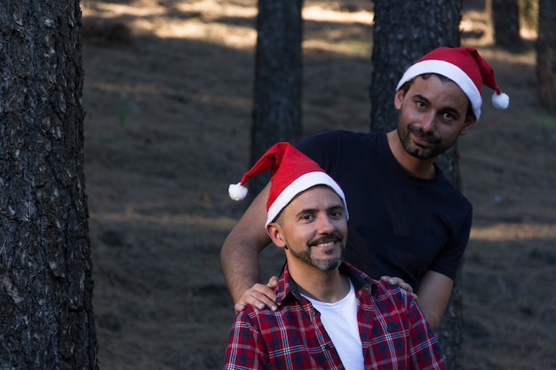 Couple gay souriant avec des chapeaux de Noël rouges posant dans un parc forestier Des hommes heureux célèbrent les vacances d'hiver