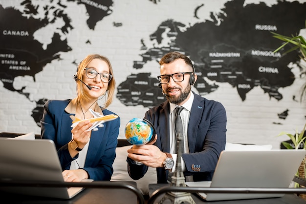 Couple de gestionnaires de voyages jouant avec un globe et un avion assis au bureau de l'agence avec une carte du monde en arrière-plan