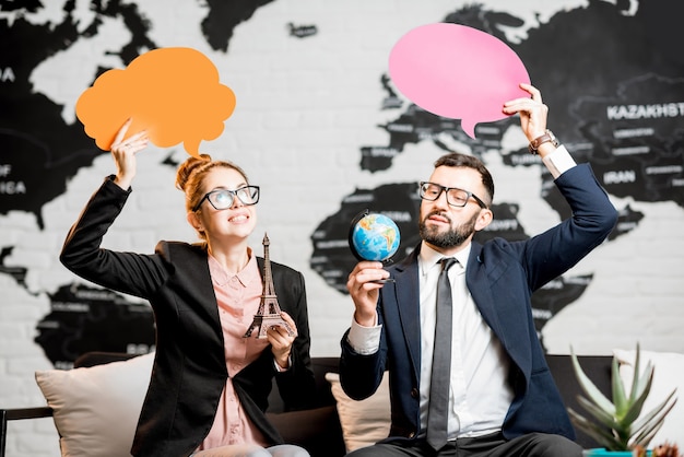 Couple de gestionnaires de voyages rêvant de bulles colorées au-dessus de la tête assis au bureau de l'agence avec la carte du monde en arrière-plan