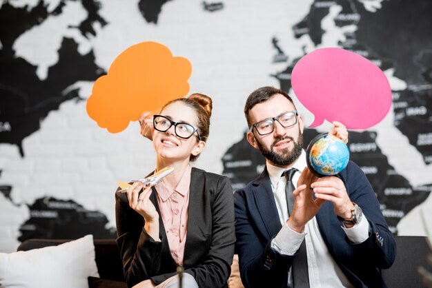 Couple de gestionnaires de voyages rêvant de bulles colorées au-dessus de la tête assis au bureau de l'agence avec la carte du monde en arrière-plan