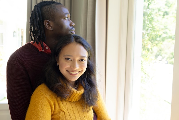 Un couple heureux et diversifié debout près de la fenêtre s'embrassant et souriant dans l'espace de copie du salon ensoleillé